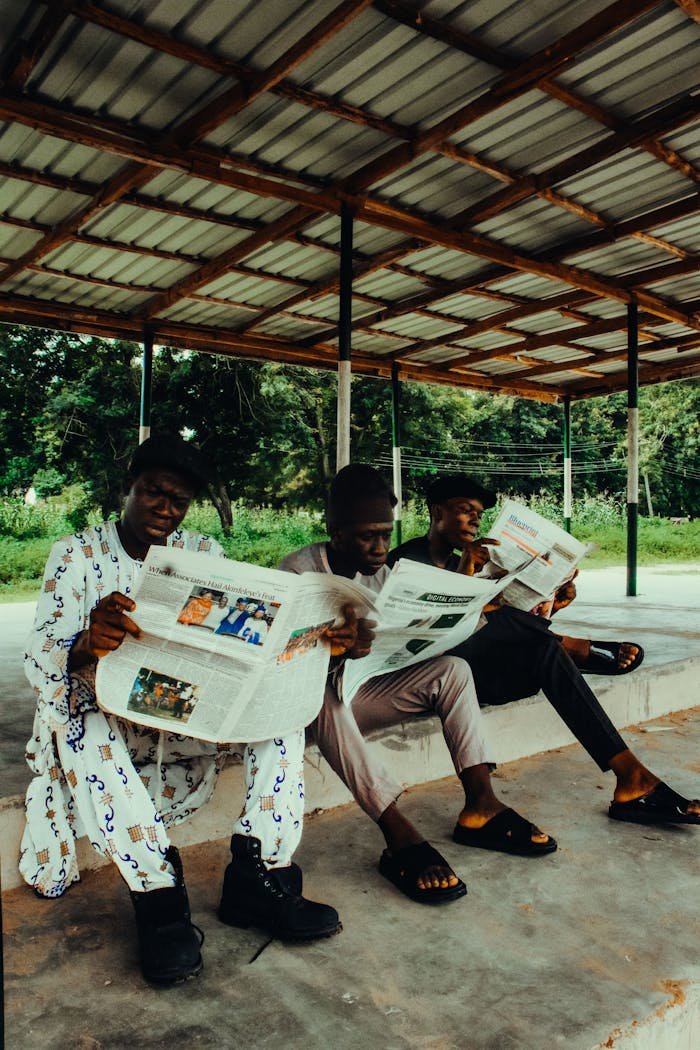 Three adults reading newspapers in an outdoor setting under a rustic shelter.